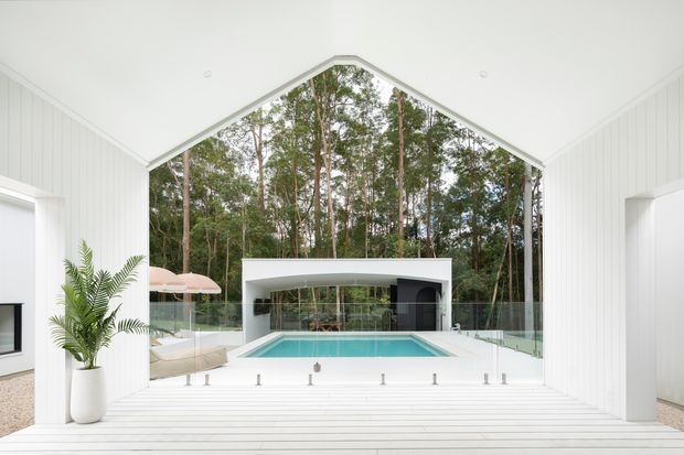 Bright, minimalist white-lined pavilion opening onto a forested outdoor pool courtyard. The room has a dramatic pitched, gabled ceiling with white vertical wall panelling and pale timber flooring, full-height glazing, and a wide deck-like threshold. A turquoise swimming pool, glass balustrade, outdoor seating, and a modern flat-roofed pool structure are visible beyond.