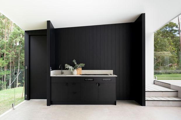 A minimalist black built-in kitchenette or bar area set against a dramatic vertical timber-panel feature wall. The space includes dark cabinetry, a pale stone benchtop, sink, tapware and a small arrangement of greenery, with full-height glazing opening to a wooded outdoor setting and steps.
