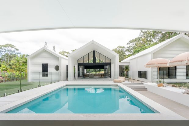 A bright contemporary residential courtyard centered on a rectangular swimming pool. White gabled buildings with weatherboard-style cladding and dark-framed glazing surround the pool, with a covered outdoor entertaining area, pale paving, and lounge furniture under a peach-toned umbrella. Lush vegetation is visible around the perimeter.