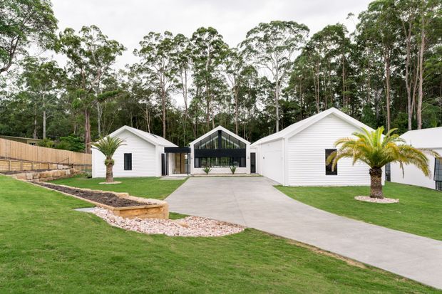 Single-storey residential property set within a landscaped lawn and eucalyptus-wooded setting. The composition features multiple white weatherboard-clad gabled wings connected by a dark-framed glazed central section, with a broad concrete driveway, palm trees, stone edging and timber boundary fencing.