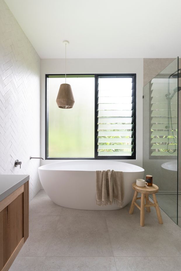 Bright contemporary bathroom featuring a freestanding white bathtub beneath a pendant light, a glass-enclosed shower, a timber vanity, large grey floor tiles, and a white textured geometric wall tile. Black-framed glazing with louvres provides natural light and ventilation.