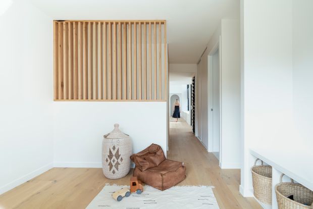 Bright, minimalist residential hallway and entry area with white walls, pale timber flooring, and a large fixed vertical timber slat screen acting as a feature partition. A low white built-in bench with woven baskets, decorative ceramic vessel, leather beanbag, and children's toys add warmth to the otherwise uncluttered space. A corridor extends toward rooms in the background.