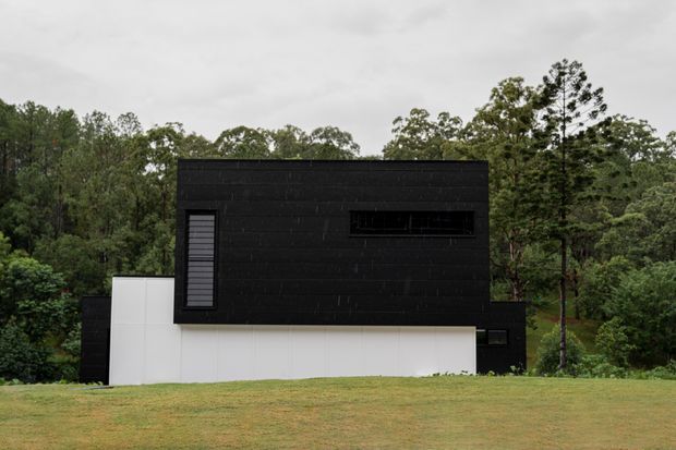 A stark modern black-and-white house set against a wooded landscape. The building has a simple rectilinear form, dark horizontal cladding across the upper volume, white rendered lower walls, and narrow dark-framed windows. A broad lawn occupies the foreground beneath an overcast sky.