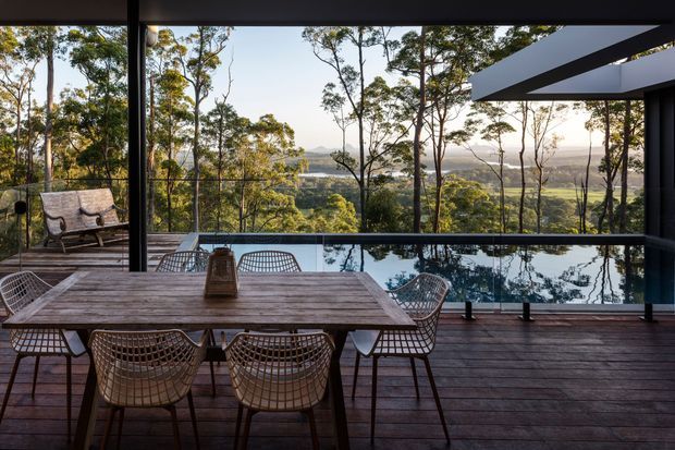 A covered outdoor dining terrace with a large timber table and woven wire chairs, overlooking a reflective pool and a wooded valley landscape. Dark structural framing, extensive glazing or open views, and timber decking create a refined connection between the home and its natural setting.