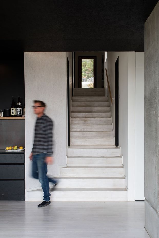 A dark, minimalist interior hallway focused on a straight stairway rising to a glazed doorway. The stair has pale wood or light stone treads and white side walls, with a simple timber handrail. A black built-in cabinet with bottles and fruit is visible at left, while tall white storage doors line the right side. The ceiling and upper walls are charcoal-toned, contrasting with the bright stair and light grey floor. A blurred person is walking through the foreground.