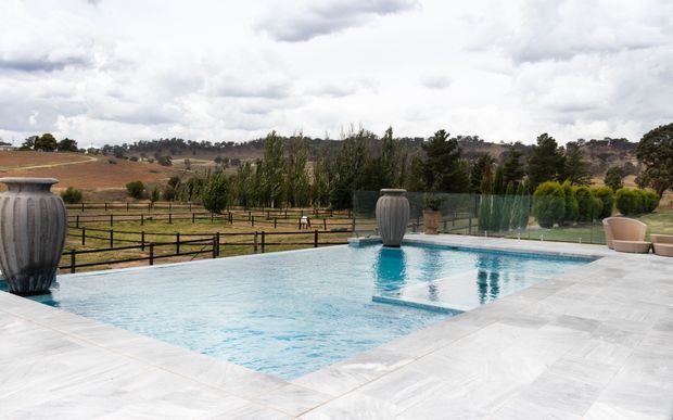 An outdoor swimming pool with pale stone paving, large decorative urns, glass pool fencing, and outdoor seating. The pool overlooks landscaped grounds and rolling rural hills under a cloudy sky.