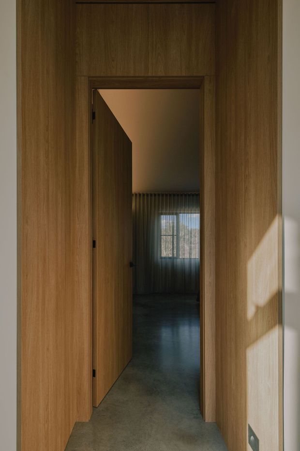 A narrow interior passage framed by warm timber wall panelling and a flush timber door opening into a dimly lit room. The adjoining room has full-height pleated curtains, a window, and a smooth grey polished-concrete-looking floor. Strong sunlight creates angular shadows across the timber and pale wall, giving the scene a quiet, refined atmosphere.