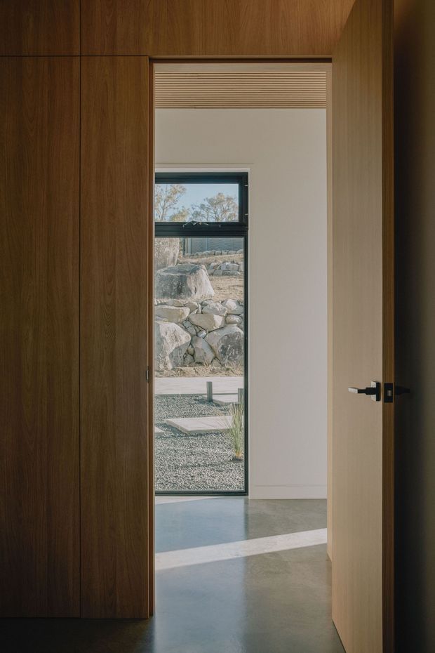 A minimalist interior view through an open timber door, showing a dark walnut-toned built-in wall or cabinetry on the left, a pale timber door on the right with a black lever handle, and a narrow black-framed window beyond. The room has smooth light-grey polished concrete flooring, white walls, and a simple light-filtering blind above the opening. Through the glazing, a rocky landscaped courtyard and gravel surface are visible. Natural sunlight creates strong diagonal shadows across the floor.