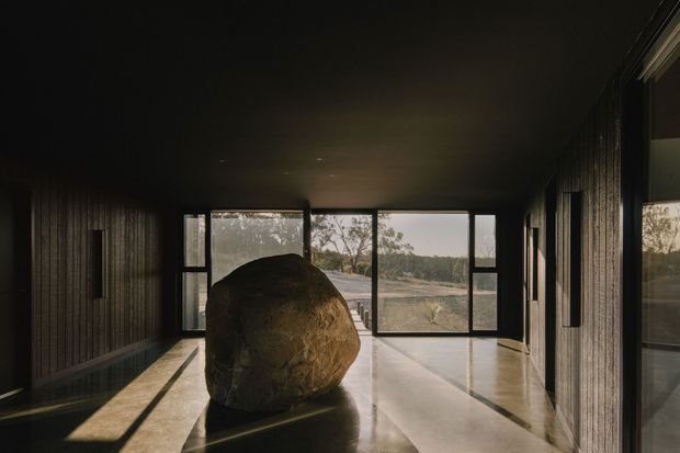 A dark, minimalist architectural interior resembling a gallery or entry hall, with polished concrete flooring, blackened or dark timber wall cladding, a low dark ceiling, and extensive floor-to-ceiling glazing. A large sculptural stone dominates the centre of the space, while the windows frame a sparse natural landscape outside.