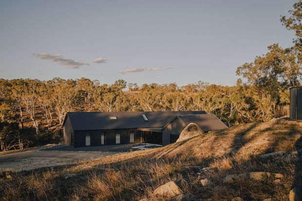 A low, dark-clad single-storey house sits within a dry, wooded landscape. The building has a long pitched metal roof with several roof windows, a projecting gabled section, and a restrained dark facade with narrow vertical windows. A gravel driveway and rocky, grass-covered foreground reinforce the rural bush setting, while warm late-afternoon light creates a subdued architectural photograph.