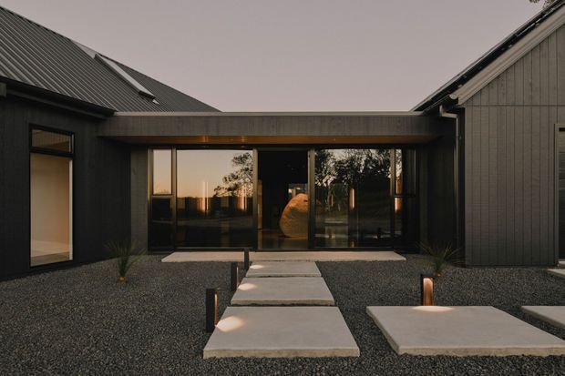 Exterior view of a contemporary dark-clad residence at dusk, centred on a glazed entry pavilion connecting two steeply pitched wings. A concrete stepping-stone path crosses a gravel forecourt toward the full-height glass doors, with restrained integrated lighting and sparse architectural planting. The composition emphasises the building facade, roof forms, and sheltered entrance.