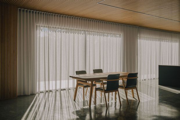 A refined contemporary dining area with a rectangular timber dining table and six timber-framed chairs positioned in front of full-height sheer curtains. The room features extensive glazing behind the curtains, a dark polished floor with strong reflected daylight, and a warm slatted timber ceiling and matching vertical timber wall treatment. A large dark display or television panel is visible at the right edge. The space is minimalist, calm, and softly lit by filtered natural light.