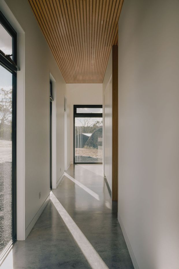 A narrow, minimalist residential hallway with polished grey concrete flooring, smooth white walls, dark-framed glazing, and a warm timber slatted ceiling. Strong sunlight creates long diagonal bands of light across the floor, while views through the glazing connect the corridor to a sparse landscaped exterior.