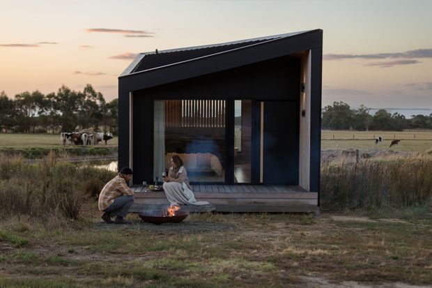 A compact dark-clad cabin or pavilion set in an open rural field at dusk. The small structure has a sharply angled single-slope roof, a glazed front facade with vertical timber screening, and a raised timber deck. Two people sit beside a small outdoor fire bowl in front of the cabin, with grazing livestock visible in the surrounding landscape.