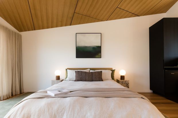 A warm, contemporary bedroom with a low-profile bed, layered neutral bedding, bedside lamps and timber bedside tables. The room features a striking timber-lined ceiling with geometric panel joints, white walls, full-height curtains, timber flooring and a large black built-in wardrobe or storage unit. A minimalist framed artwork hangs above the bed.