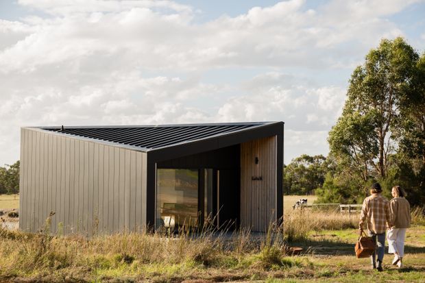 A compact modern pavilion or small dwelling set in an open rural landscape. The building has dark vertical cladding, a low mono-pitched roof with exposed dark roof ribs, a deep black-framed recessed entry, and warm timber-lined surfaces around the doorway. Two people approach along a rough grass path, with mature trees and open fields behind.