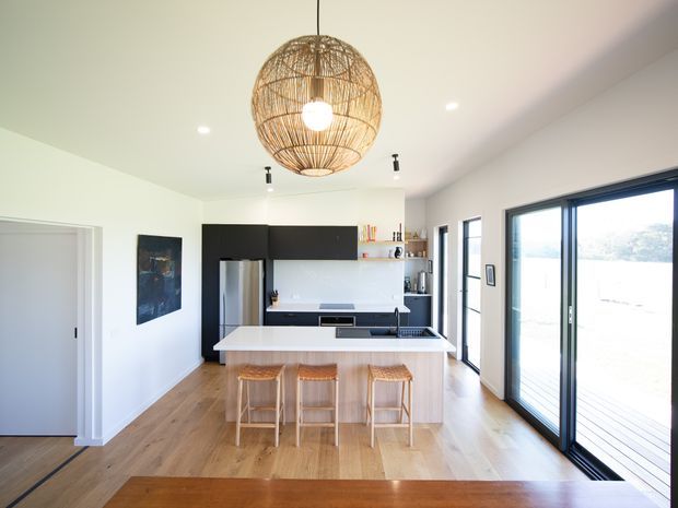 Bright contemporary open-plan kitchen viewed from the adjoining living or dining area. The room features pale timber flooring, a white island with three timber bar stools, flat-panel black cabinetry, white benchtops and splashback, integrated appliances, large black-framed glazed doors, and a woven pendant light. A deck or outdoor area is visible beyond the glazing.