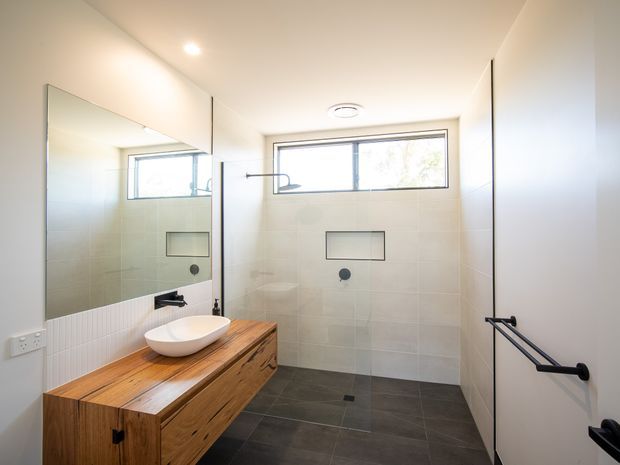 Bright contemporary bathroom with a walk-in shower, frameless glass screen, large wall mirror, pale tiled walls, dark tiled floor, and a timber vanity with a white vessel basin. A horizontal window provides natural light, while black fixtures and grab rails add contrast.