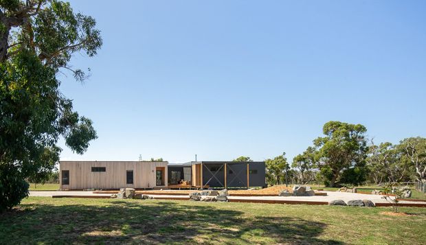 Wide exterior view of a low-profile single-storey contemporary home set in a spacious rural or semi-rural landscape. The building has a long horizontal composition, timber-clad walls, dark contrasting wall sections, large glazed openings, and a flat or very low-pitched roof. Native-looking trees, lawn, exposed rocks, and landscaped garden beds surround the house under a clear blue sky.