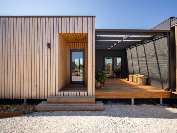 Exterior view of a compact contemporary timber-clad dwelling with a recessed entry and covered timber deck or verandah. Vertical light-wood cladding, dark-framed glazing, a pergola-like roof structure, and screened outdoor living area are clearly visible.