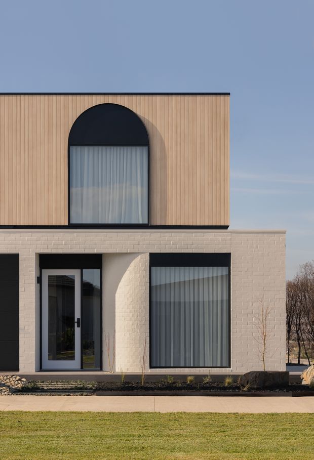 Front exterior view of a contemporary two-storey house with a rectilinear form, pale painted brick lower level, vertically lined warm timber-clad upper level, and bold black-framed glazing. A large arched upper window and dark-framed ground-floor windows and entry door create strong geometric contrast. Minimal landscaping and a lawn sit in front of the facade.