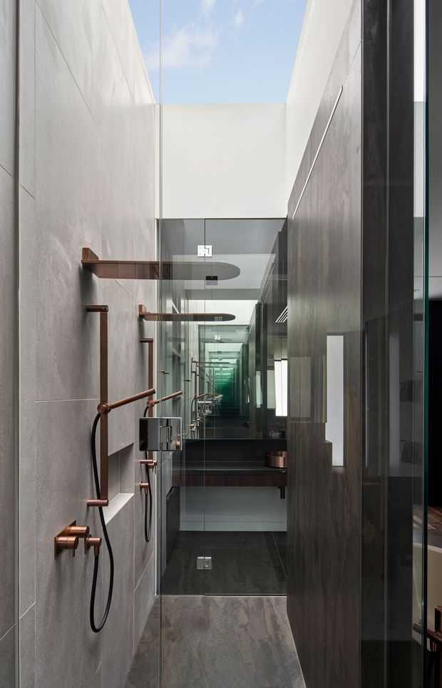 A narrow, vertically proportioned contemporary bathroom with a walk-in shower, dark stone-look wall and floor finishes, large mirrors creating repeated reflections, and exposed shower fittings in a warm copper or bronze finish. A glazed opening above brings in daylight and blue sky, while dark timber or wood-look elements frame the space.