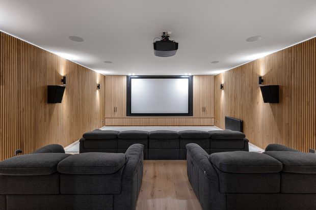 A dedicated home cinema/media room viewed from the rear seating area, with multiple rows of dark sectional seating facing a large projection screen. The room features warm vertical timber slat wall panelling, integrated wall sconces, concealed ceiling lighting, recessed downlights, a ceiling-mounted projector, and timber flooring. The atmosphere is dark, comfortable, and cinematic.