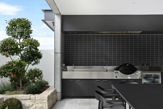 A covered outdoor kitchen and dining area with dark built-in cabinetry, a stainless-steel barbecue, and a black tiled splashback. A dark dining table and chairs occupy the foreground, while a landscaped planter with a small tree borders the space. The design has a crisp contemporary appearance with strong black, grey, and stainless-steel finishes.