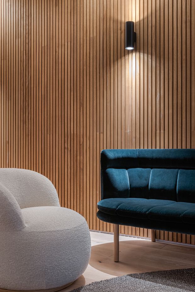 A warmly lit interior seating area featuring a full-height vertical timber slat feature wall, a sculptural cream boucle lounge chair, and a deep teal velvet upholstered sofa with slender timber legs. A black cylindrical wall light casts an upward and downward glow across the timber wall, while a timber floor and dark textured rug complete the refined, contemporary setting.