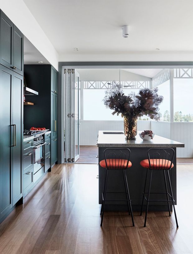 A bright contemporary kitchen with floor-to-ceiling dark teal cabinetry, integrated appliances, a central island with a light stone benchtop, and two black bar stools with red upholstered seats. The kitchen opens directly to a glazed outdoor area with timber decking and decorative structural framing. Warm timber flooring, white walls, and abundant natural light create a refined, airy interior.