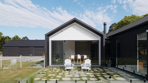 Exterior view of a contemporary dark-clad residential building with a central white gabled pavilion opening onto a paved courtyard. The courtyard includes a grid of large square pavers set among gravel and grass, outdoor seating, low planting, and a transparent boundary fence. Large glazed openings and dark vertical cladding define the adjoining wing, while a matching dark outbuilding sits to the left.