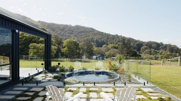 A landscaped outdoor entertaining area viewed from beside a dark-clad contemporary building, featuring a small rounded swimming pool, glass safety fencing, a paved stepping-stone terrace with Adirondack-style chairs, and a broad grassy setting backed by wooded hills. The building has dark vertical cladding, extensive glazing, and a sloping roof.