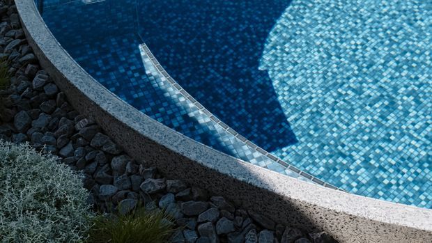 Close-up exterior detail of a curved swimming pool with blue mosaic tile lining, a pale textured concrete or stone coping edge, and dark rock landscaping with sparse planting.