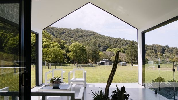 A bright contemporary interior opening onto a rural landscape through expansive black-framed glazing. The room has a dramatic triangular, house-shaped glazed opening, pale flooring and walls, and a small white outdoor-style seating arrangement with potted plants. A glass balustrade or pool-like reflective surface is visible at the right, while wooded hills and open grassland appear beyond the building.