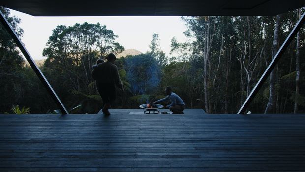 A dark, expansive timber deck or covered outdoor platform framed by dramatic angled structural supports, opening toward a wooded landscape and distant hills. Two people sit near a small low fire or cooking feature, creating a secluded, nature-connected outdoor gathering space. The image has a cinematic, moody atmosphere with strong contrast between the shadowed structure and bright forest outlook.