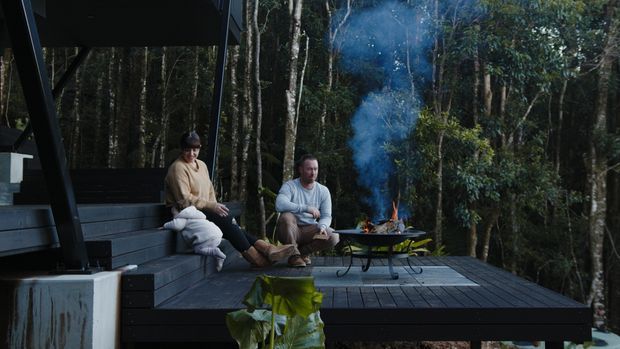 A dark timber outdoor deck platform set within dense forest, with tiered built-in bench seating beneath a simple roof or canopy. Two people sit around a freestanding fire bowl, creating a secluded woodland gathering space. The deck structure and black timber finishes dominate the image, with smoke rising into the trees.