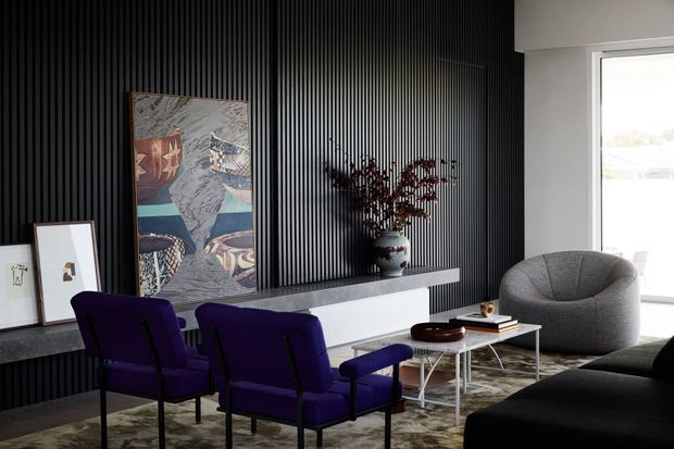 A refined contemporary living room with a dramatic black vertical slatted feature wall, white floating media console, and grey stone-look ledge. Two deep purple lounge chairs face the seating area, accompanied by a sculptural grey armchair, low white-framed coffee table, and dark sofa. Abstract artwork, a vase of branches, and a patterned rug add texture and colour. Bright glazing at right provides natural light.
