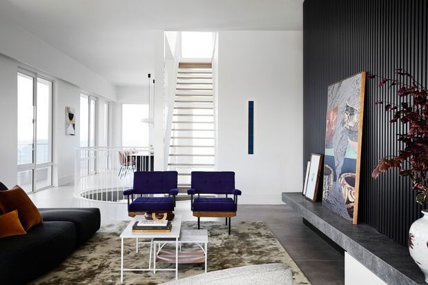 Bright contemporary living room with a black sofa, two purple lounge chairs, layered rugs, and a white coffee table. A dramatic black vertical-slat feature wall with a long dark stone hearth, artwork, and decorative objects forms the main focal point. White walls, extensive glazing, and an open stair with timber treads create an airy, multi-level interior.