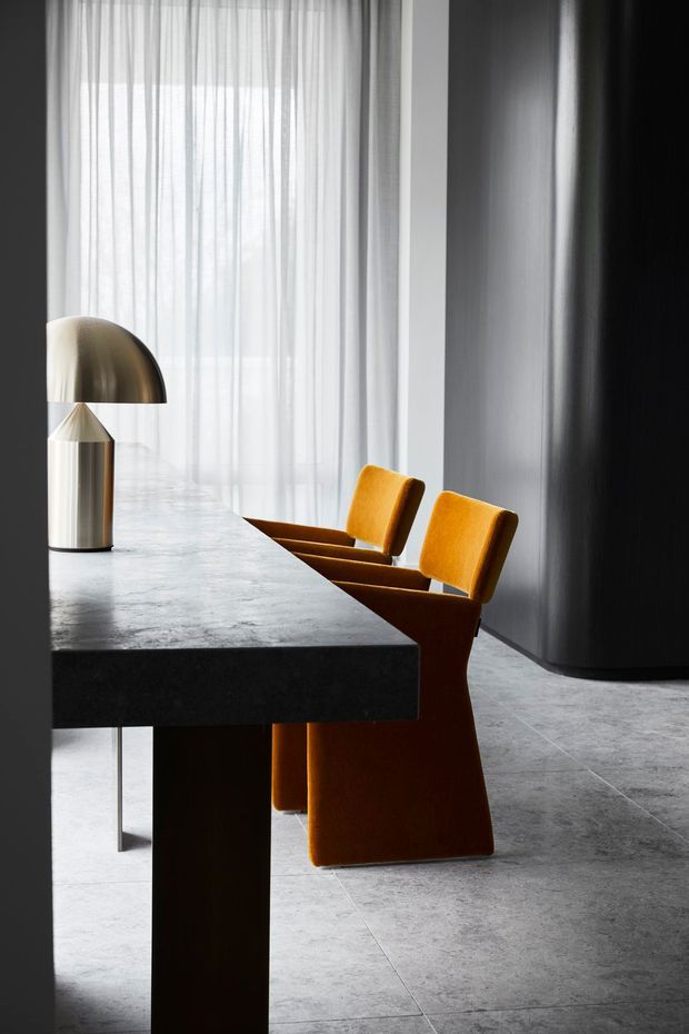 A sophisticated contemporary dining area with a long dark stone or concrete dining table, sculptural ochre upholstered dining chairs, and a metallic table lamp. Floor-to-ceiling sheer curtains provide soft daylight, while dark walls and large grey stone-look floor tiles create a dramatic, refined interior.
