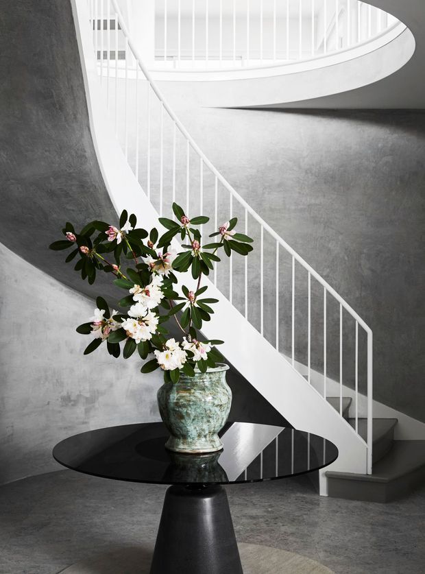 A sculptural curved staircase with slim white vertical balustrades winds through a tall, grey textured interior. A black pedestal table with a glossy round top and a large weathered ceramic vase of flowering branches occupies the foreground. The space has a dramatic monochrome palette and strong curved architectural forms.