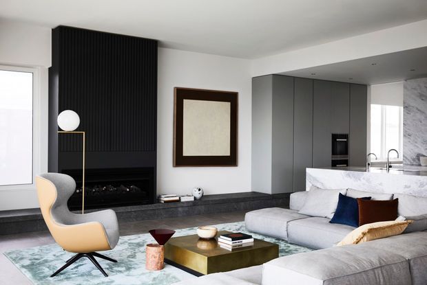 A refined open-plan living room with a sculptural two-tone lounge chair, large pale sectional sofa, low brass-toned coffee table, and a black linear fireplace set into a dramatic vertically ribbed feature wall. The adjoining kitchen has dark grey full-height cabinetry, a white marble or stone island, and marble wall cladding. The room uses a restrained neutral palette with dark architectural accents and contemporary artwork.