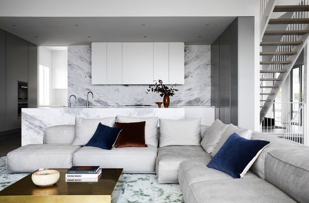 A sophisticated open-plan living room with a large pale sectional sofa facing a low coffee table, positioned in front of a marble kitchen island and full-height marble splashback. White flat-panel cabinetry, integrated dark cabinetry, recessed ceiling lights, and a contemporary open-riser staircase with glass balustrade create a clean, refined interior.