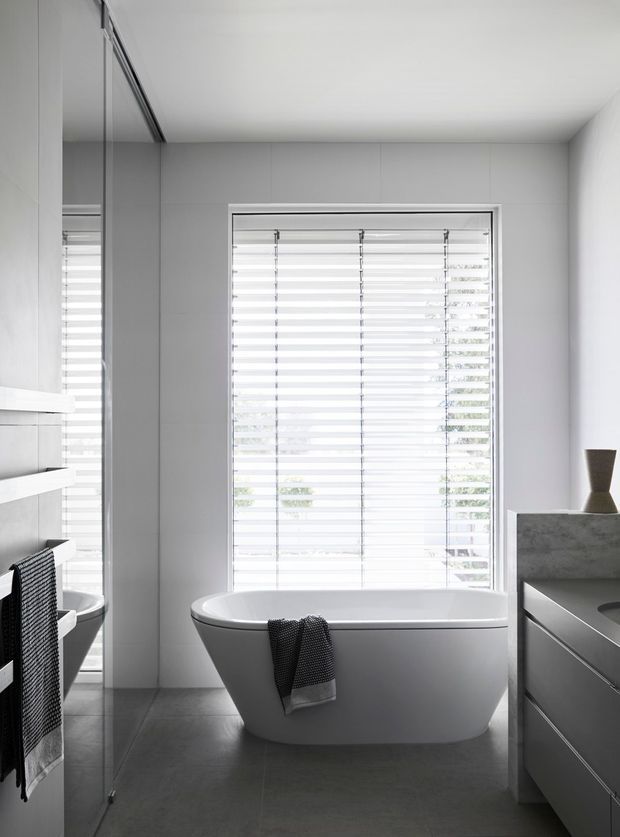 A bright, contemporary bathroom featuring a freestanding white bathtub positioned beneath a large window with horizontal blinds. The room has pale grey wall panels, dark grey floor tiles, mirrored cabinetry on the left, and a grey vanity with a stone-like benchtop on the right. The overall composition is clean, restrained, and monochromatic.
