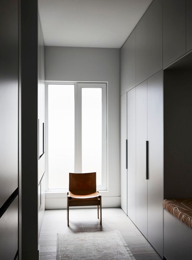 A narrow, minimalist dressing room or wardrobe passage with full-height matte grey built-in cabinetry on both sides, a large frosted or overexposed window at the end, pale flooring, and a simple brown leather chair. A built-in upholstered bench is visible on the right, with a neutral rug adding softness to the otherwise highly restrained space.