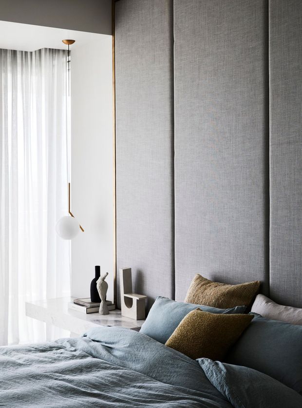 A refined contemporary bedroom with a tall upholstered grey feature wall behind the bed, layered blue-grey and ochre bedding, and a slim white stone bedside ledge. Sheer full-height curtains filter daylight beside a sculptural pendant light, while minimalist decorative objects sit on the ledge.