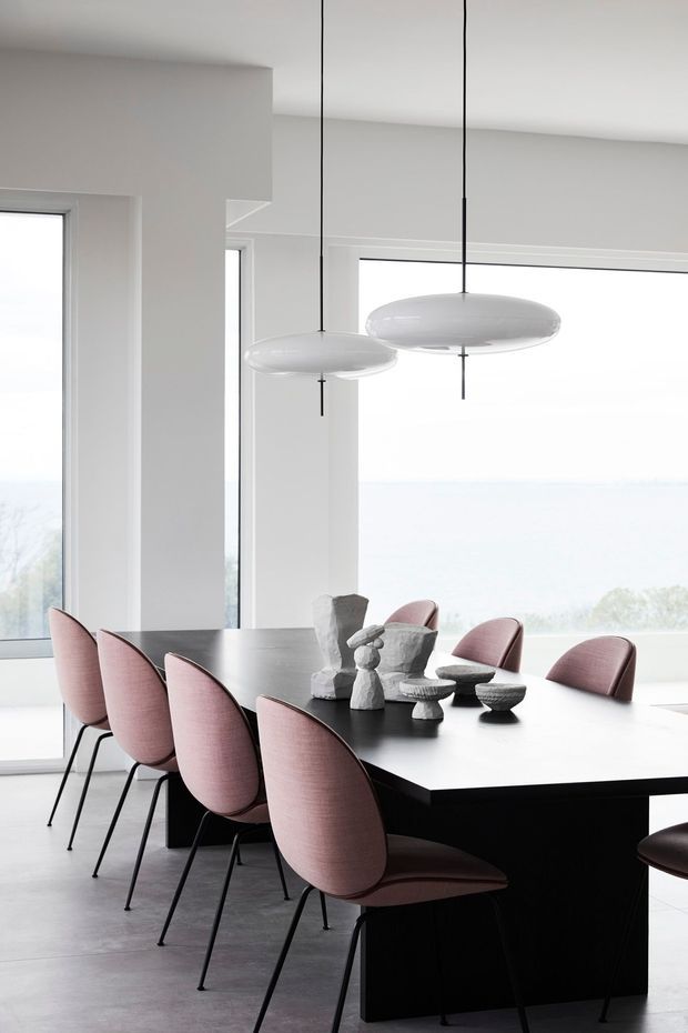 A minimalist dining area with a long black rectangular dining table surrounded by blush-pink upholstered chairs with slender black legs. Sculptural monochrome objects form a centrepiece, while two suspended white disc pendant lights hang above. Full-height windows provide abundant natural light and a pale exterior view; the room has white walls, a smooth light-grey floor, and a clean contemporary fit-out.