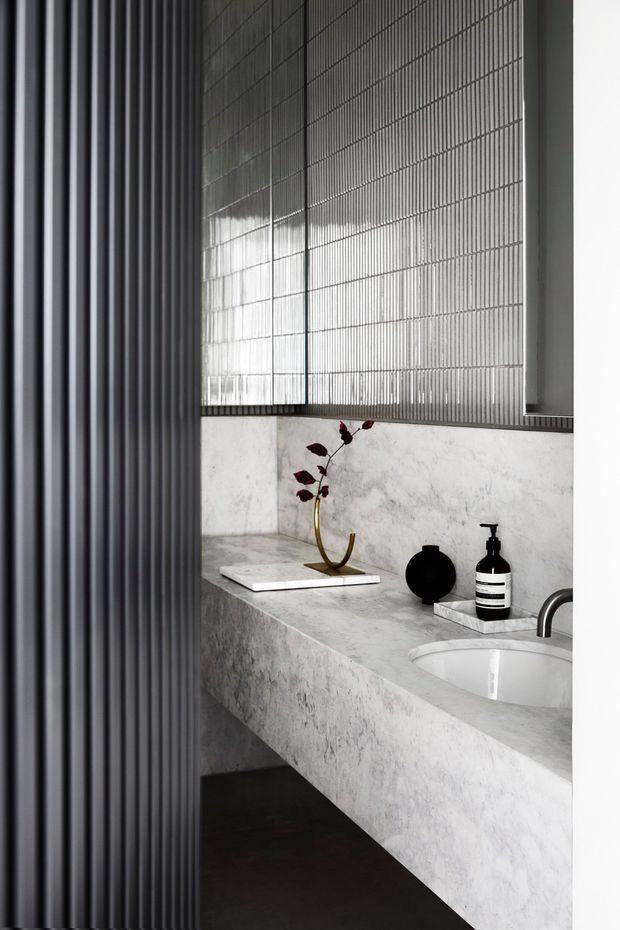 A refined minimalist bathroom vanity featuring a monolithic pale veined stone benchtop and integrated basin, dark curved tapware, a ribbed translucent glass window, and a vertical dark fluted screen or wall panel. Black decorative accessories provide contrast against the light stone surfaces.