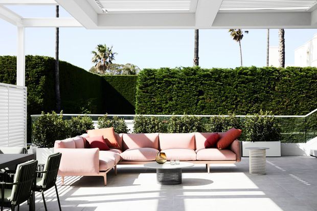 Bright contemporary outdoor lounge terrace beneath a white slatted pergola, furnished with a large blush-pink sectional sofa, dark dining chairs, and low coffee tables. The terrace has pale tiled paving, white planters, glass balustrades, and dense clipped hedging with palm trees beyond.