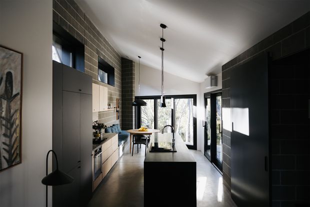 A narrow, contemporary galley-style kitchen with a central black island, dark integrated cabinetry, pale timber cabinetry, and a light stone or concrete-look floor. Dark rectangular wall tiles wrap the upper walls, while full-height glazing and a glazed door bring in strong natural light. Black pendant lights, a modern gooseneck tap, and minimalist appliances contribute to the refined monochromatic scheme.
