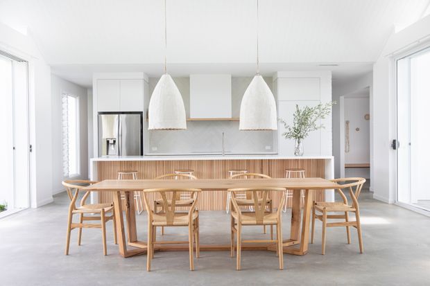 Bright open-plan kitchen and dining area with a long light-wood dining table surrounded by timber wishbone-style chairs. The kitchen features white flat-panel cabinetry, stainless-steel appliances, a pale stone or composite benchtop, and a vertically slatted timber island front. Two oversized white pendant lights hang above the table, with large glazed doors and windows contributing to an airy, minimalist coastal feel.
